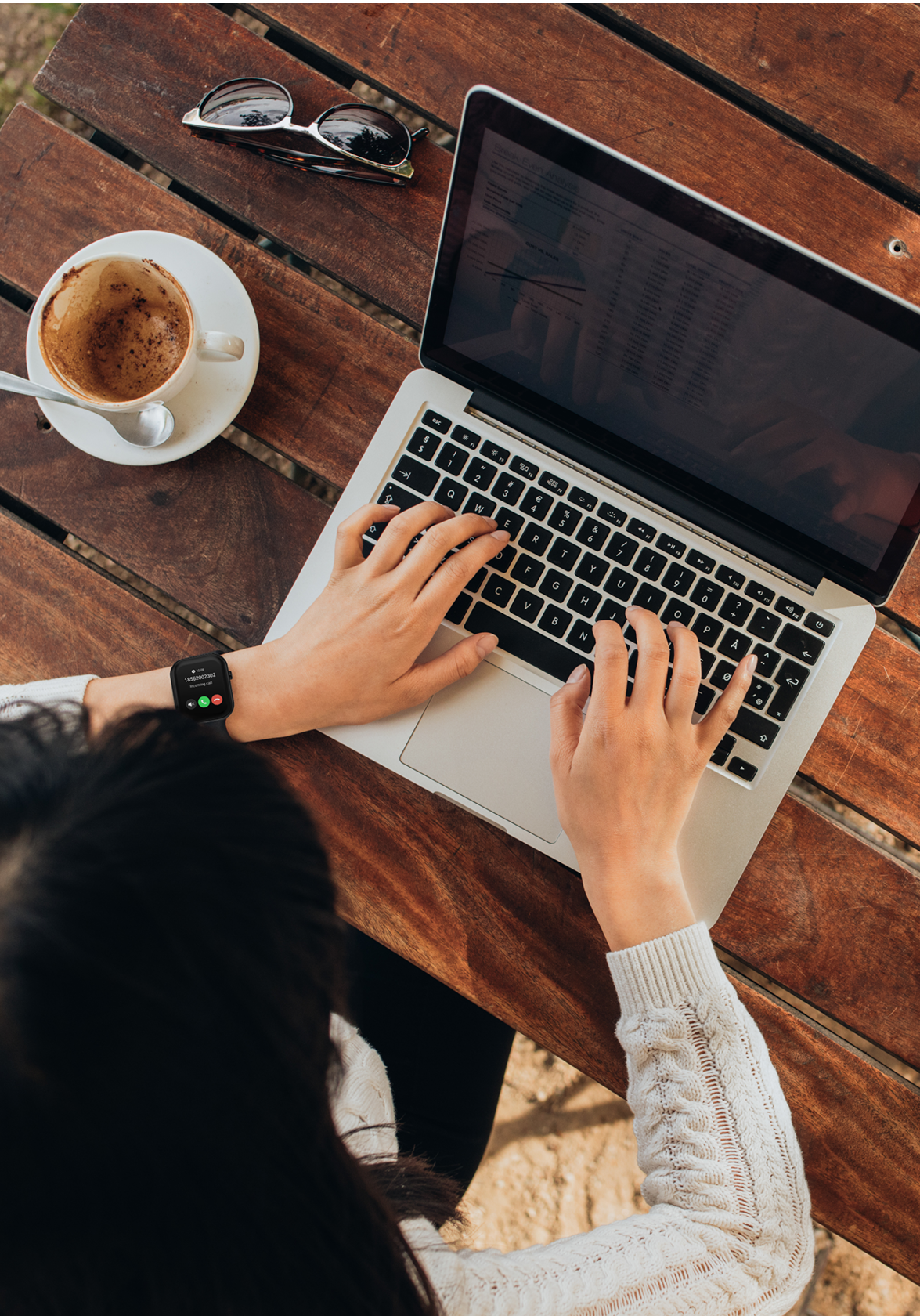Person using a laptop on a wooden table with a cup of coffee and sunglasses.